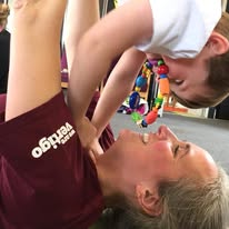 A joyful interaction between a smiling adult and a child during a playful activity, both positioned on a carpeted surface.