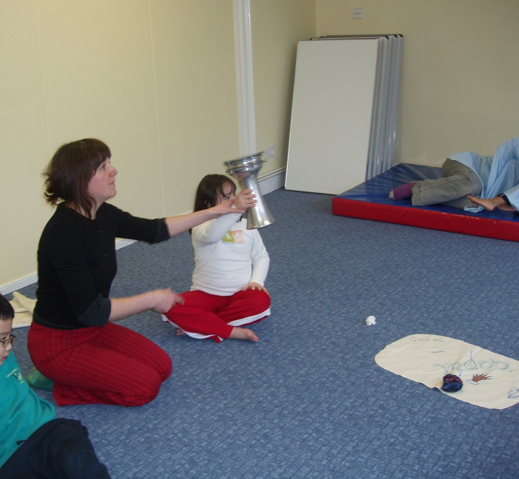 A group of children participating in a dramatherapy session, with one child holding a silver drum while sitting on the floor. An adult facilitator is engaging with the children, and others are seated or lying in the background.