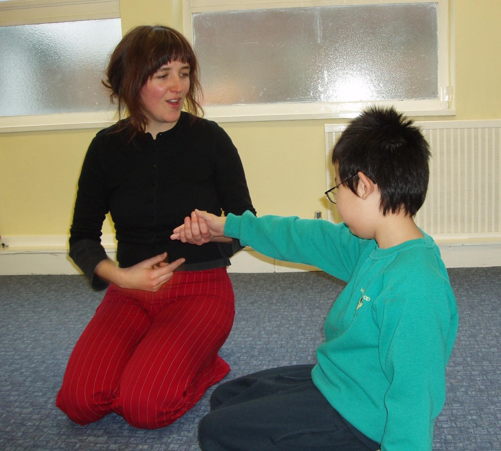 A woman with dark hair and a black shirt is sitting on the floor, engaging with a young boy wearing a turquoise sweatshirt. They are both seated, with the boy extending his arm while the woman holds his hand, smiling.