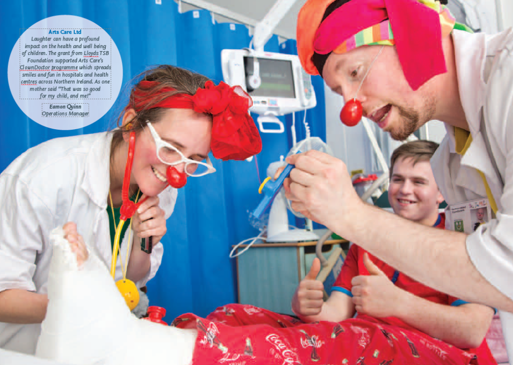 Two clown doctors engaging with a child in a hospital setting, one wearing a red clown nose and glasses, while the other holds a toy. A young adult smiles in the background.