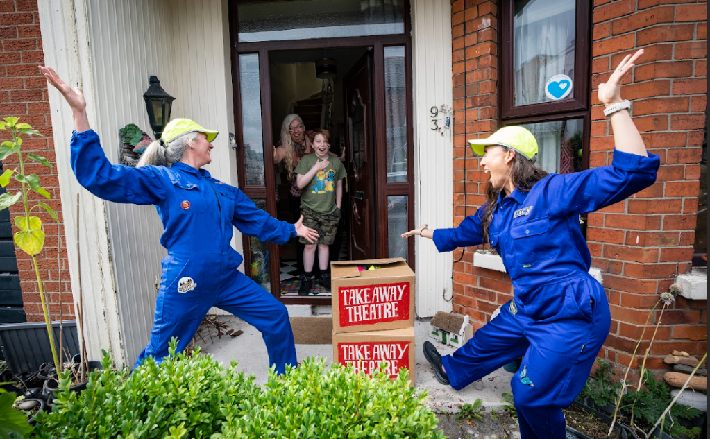 Two performers in blue overalls and caps joyfully pose outside a house while a child watches from the doorstep, with takeaway boxes labeled 'Take Away Theatre' in front.